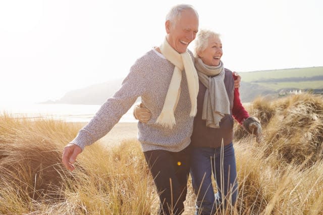 Senior Couple Walking Through Sand Dunes On Winter Beach Smiling