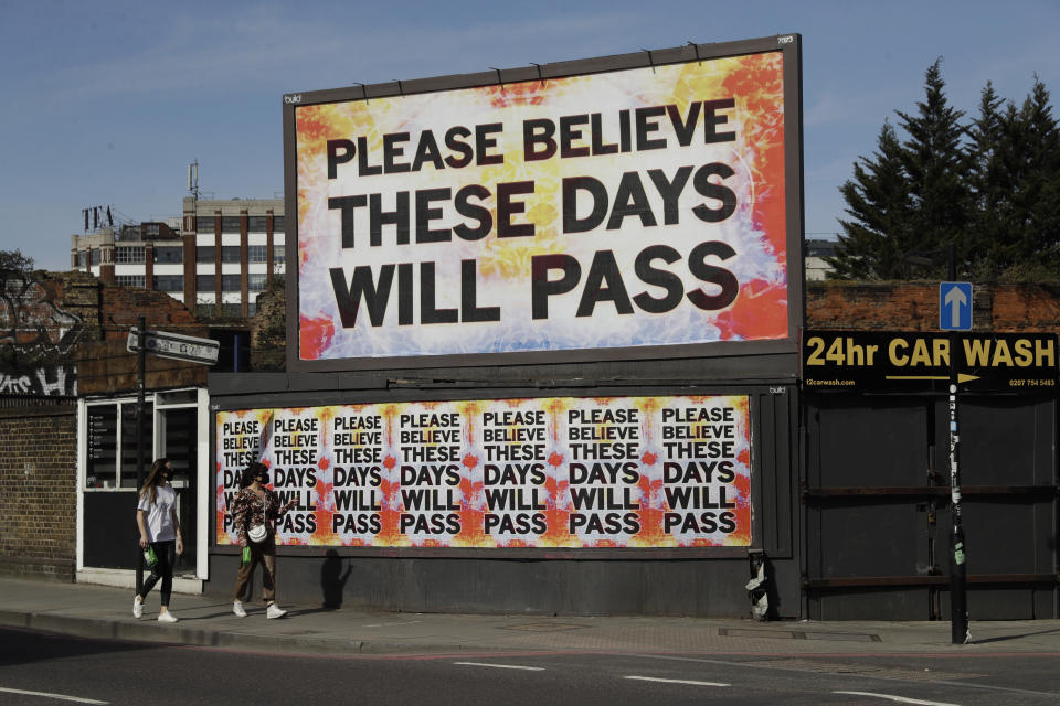 Women wearing face masks to protect from coronavirus walk past a billboard with a work by artist Mark Titchner in east London, Tuesday, April 7, 2020. The new coronavirus causes mild or moderate symptoms for most people, but for some, especially older adults and people with existing health problems, it can cause more severe illness or death. (AP Photo/Matt Dunham)