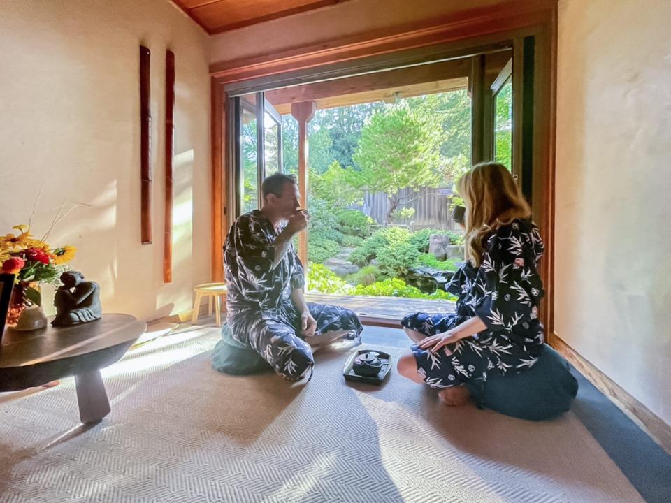 Two people sit on meditation pillows sipping from mugs in a white-walled room. Behind them is a floor-to-ceiling window with lush greenery in sight behind it.