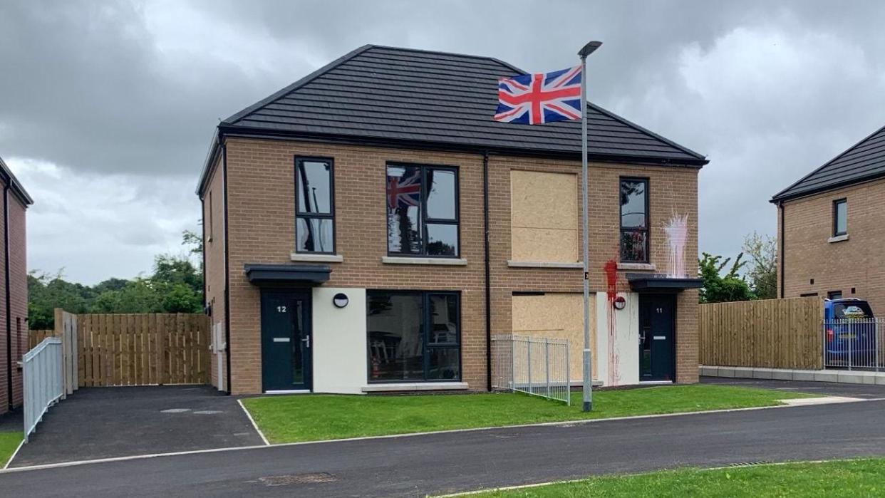 An image of the damaged home alongside other homes in the estate. A Union Jack flag is flying on a lamppost outside the house. 