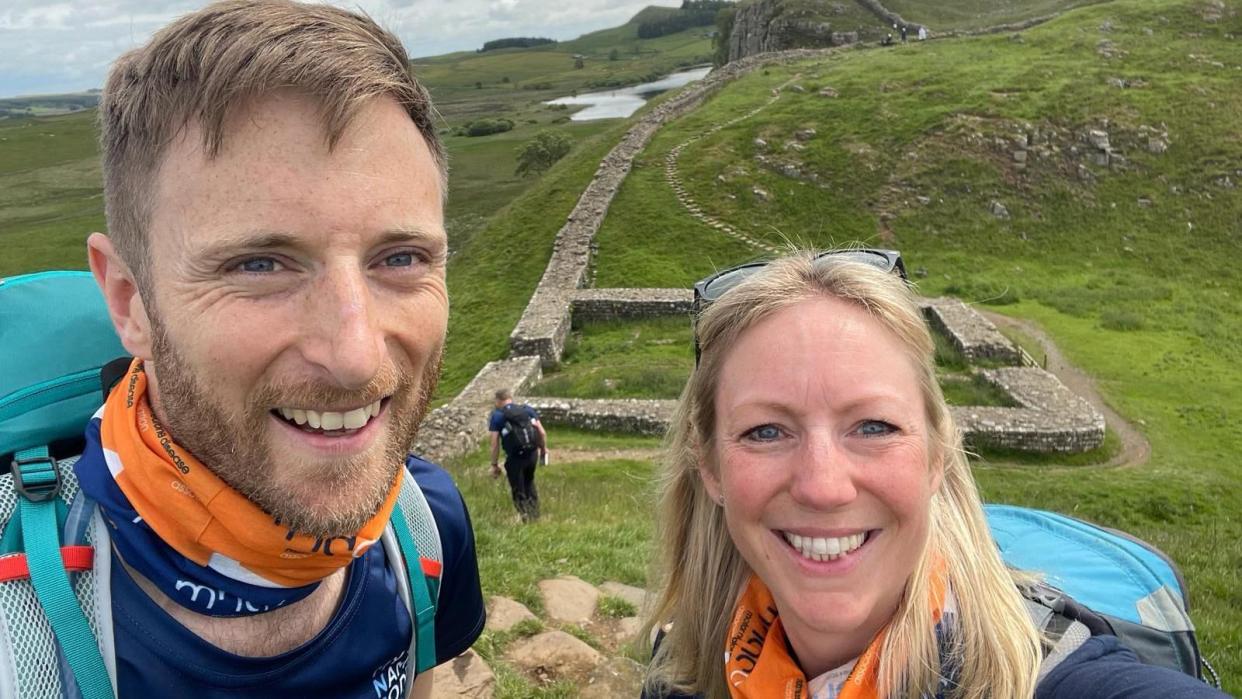 Luke Hames-Brown has a mousey brown beard and short hair and Kate has blonde, shoulder length hair. They're both carrying rucksacks and MND Association snoods 