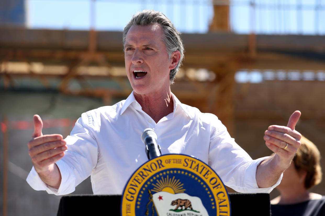 Gavin Newsom, in a white button-down shirt with rolled-up sleeves, stands at a podium with a single microphone and a seal that reads: the Governor of the State of California.