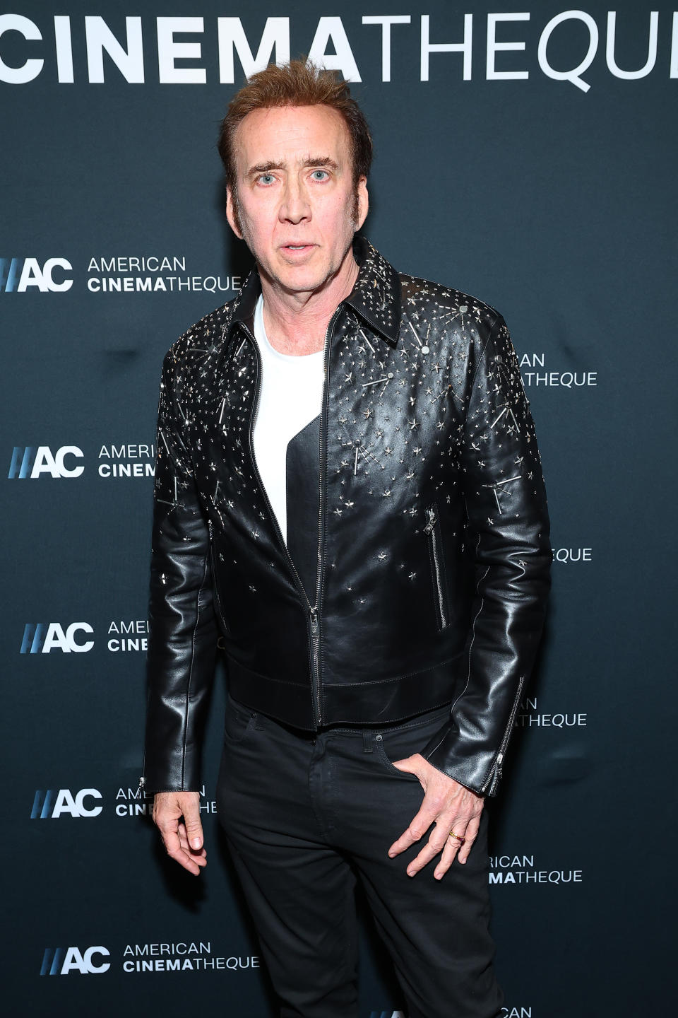 Nicolas Cage at an American Cinematheque event, wearing a studded leather jacket over a white shirt. Step and repeat backdrop with event logo in the background