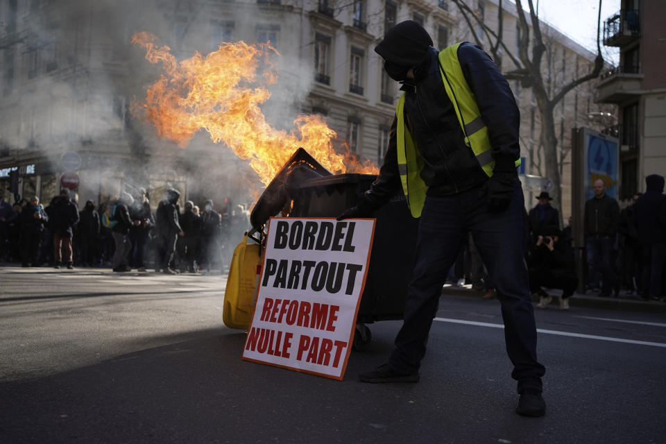 A demonstrator places a placard near a burning garbage bin reading : "Mess Everywhere, Reform Nowhere" during a march against pension reforms in Lyon, central France, Tuesday, Feb. 7, 2023. Public transportation, schools and electricity, oil and gas supplies were disrupted on Tuesday in France as demonstrators are taking to the streets for a third round of nationwide strikes and protests against the government's pension reform plans. (AP Photo/Laurent Cipriani)