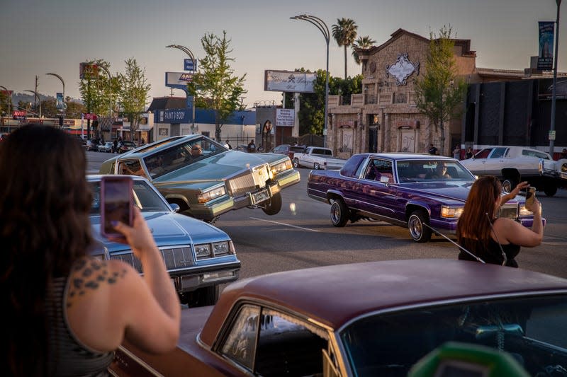 Two women recording a low rider cruise with two low low Cadillac Coupe DeVilles driving on Van Nuys Blvd. in Los Angeles