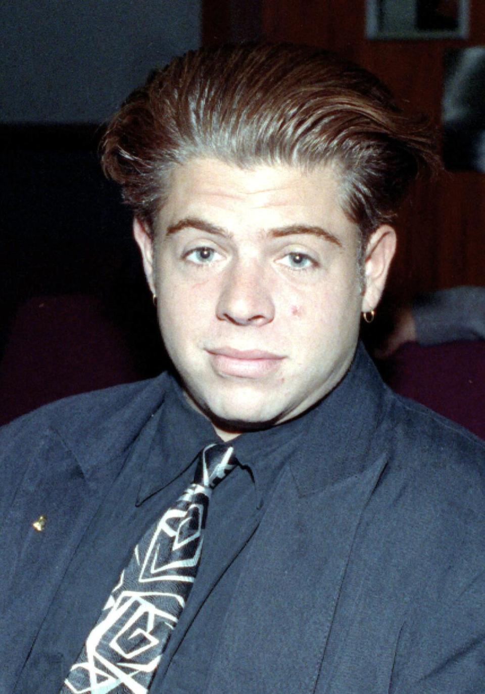 A man with slicked back brown hair and blue eyes wearing a collared shirt and a patterned necktie
