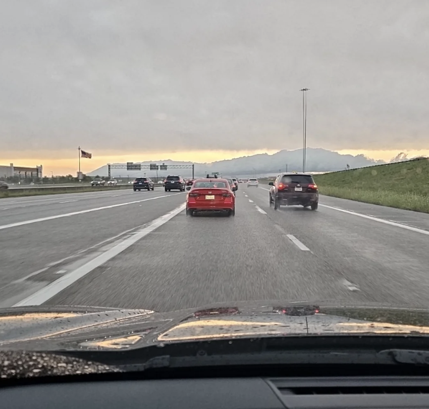 Cars driving on a multi-lane highway with road signs visible in the distance and mountains on the horizon during a cloudy sunset