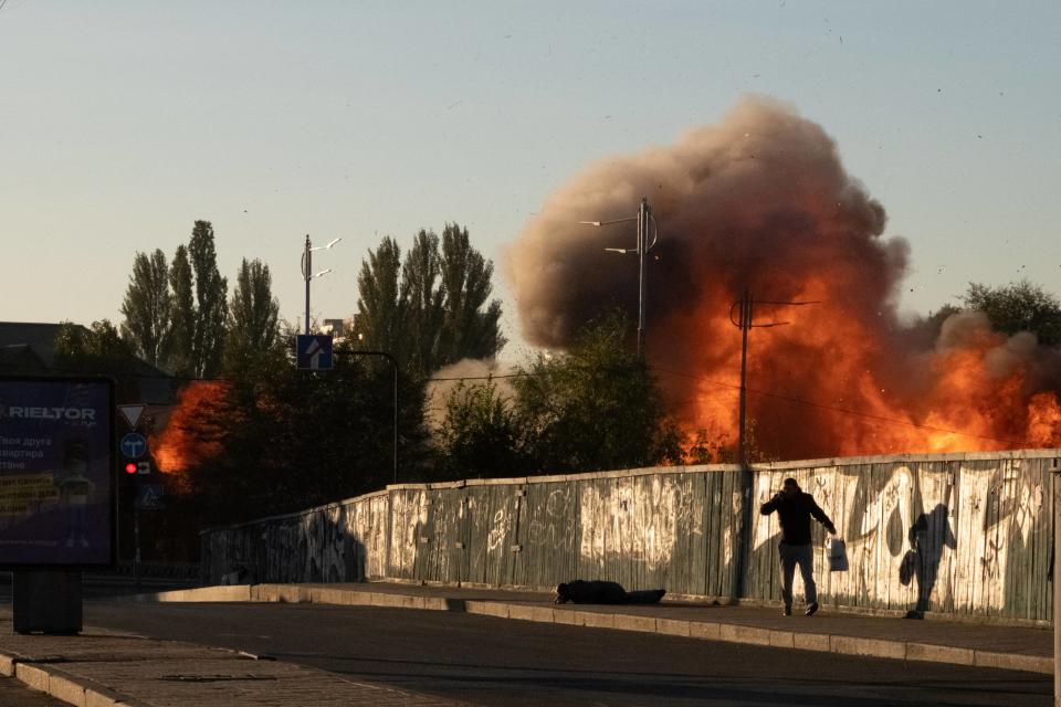 Two men, one fallen on the ground, are seen after a blast. High flames and smoke are in the background.