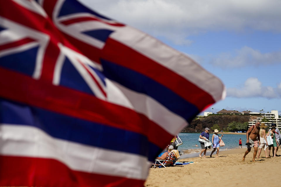 Tourists walk on Kaanapali Beach in front of a flag of Hawaii planted in the sand, Wednesday, Dec. 6, 2023, in Lahaina, Hawaii. Residents and survivors still dealing with the aftermath of the August wildfires in Lahaina have mixed feelings as tourists begin to return to the west side of Maui. (AP Photo/Lindsey Wasson)