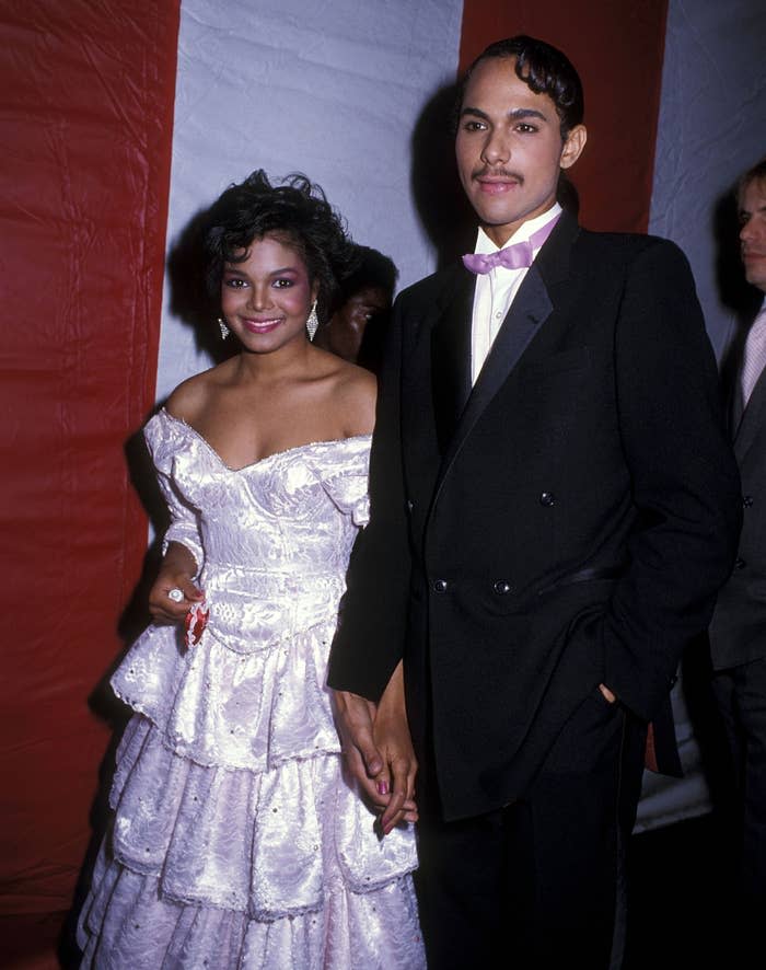 Janet Jackson and a companion hold hands on the red carpet. Janet wears a strapless ruffled dress and her companion wears a tuxedo with a pink bow tie.