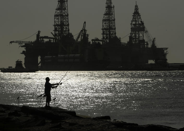 Silhouette of a man fishing off a coast near docked oil drilling platforms.
