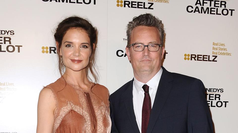 katie holmes and matthew perry pose for a photo each with one arm around the other while standing in front of a white background, she wears a rose sleeveless dress and holds a gold clutch, he wears a blue suit, red tie and white collared shirt with black rimmed glasses