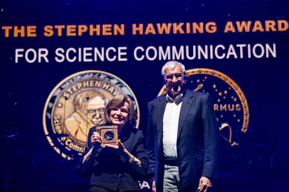 two people stand on stage and smile, the one on the left is holding an award.