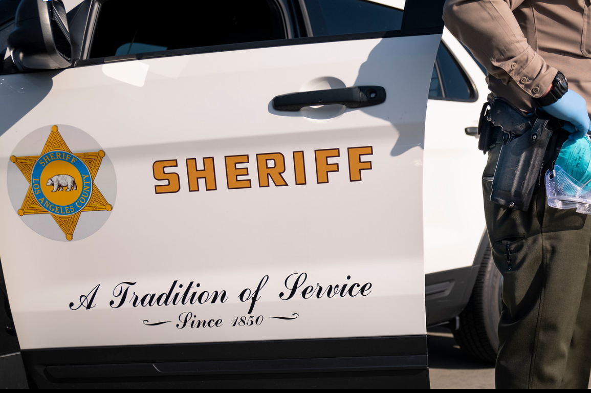 A Los Angeles County Sheriff's Office armed deputy standing next to a patrol car. A 17-year-old girl reportedly grabbed a deputies holstered firearm on March 24, 2024 and allegedly fatally shot herself inside the lobby of it's Industry station.