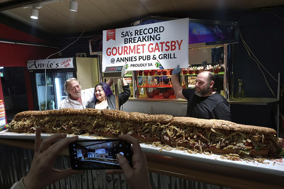 In this photo provided by Joe Redelinghuys. left, Annie Redelinghuys, centre, and chef Michele Steyn, right, pose for a photograph at Annies Ladies Bar, with a giant Gatsby sandwich on display at the establishment in Cape Town, South Africa, Saturday, June 29, 2024. A South African restaurant and bar has served up a 3-meter-long (9.8 feet) sandwich in the latest offering from an establishment with a taste for the supersize. (Annies Ladies Bar via AP)