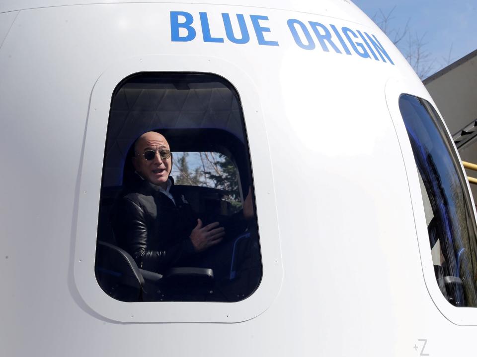 jeff bezos bald wearing sunglasses gestures with open mouth in the window of a blue origin space capsule