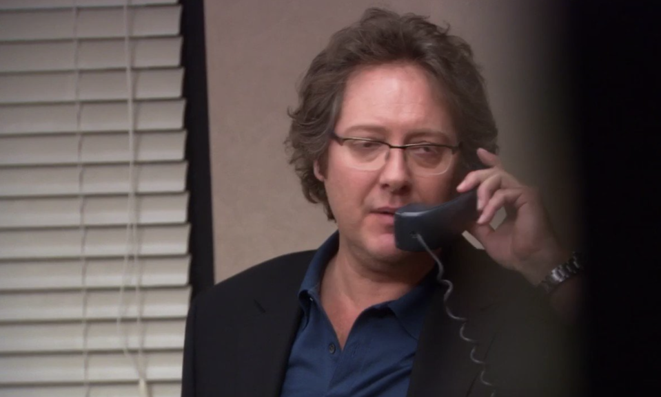 Man portraying a character on a TV show, speaking on a phone in an office setting