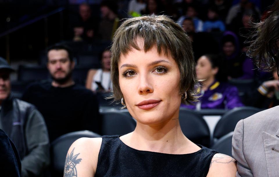 Woman with short hair and tattoos wearing a tank top and jeans, holding a glass, at a sports event