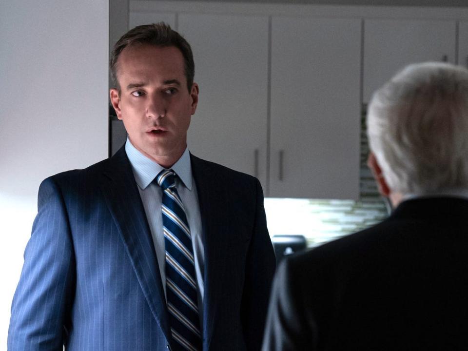 Two tall white men in suits facing a shorter, older man in an office break room.