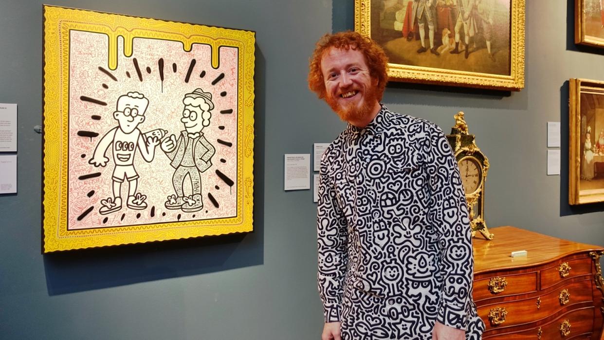 The artist Mr Doodle, a man with ginger hair and wearing a doodle-covered shirt and trousers, stands in front of a canvas of his work showing two figures with lots of expressive lines and a painted frame. He is in an art gallery. 