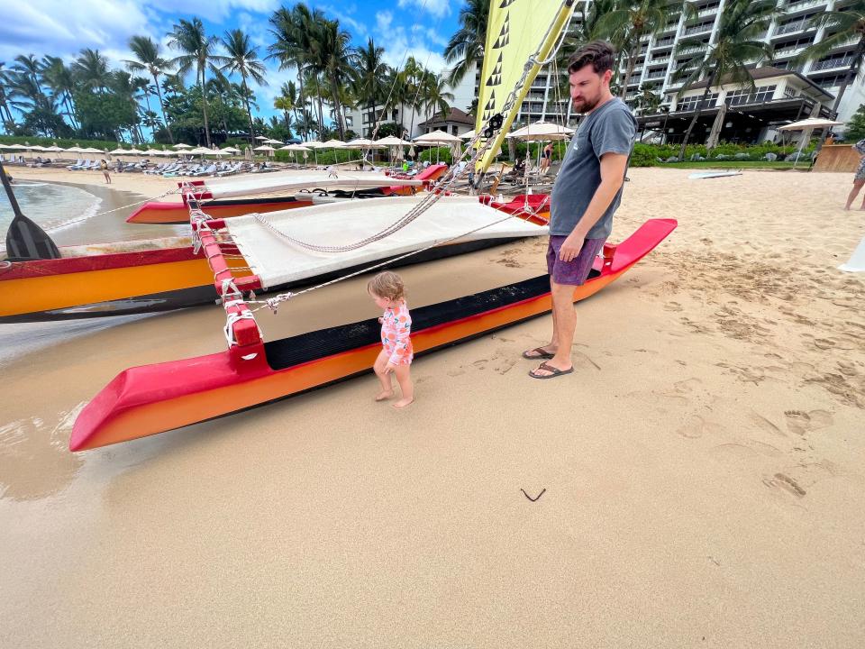 A baby walks next to a catamaran-style sailing boat with her father watching.