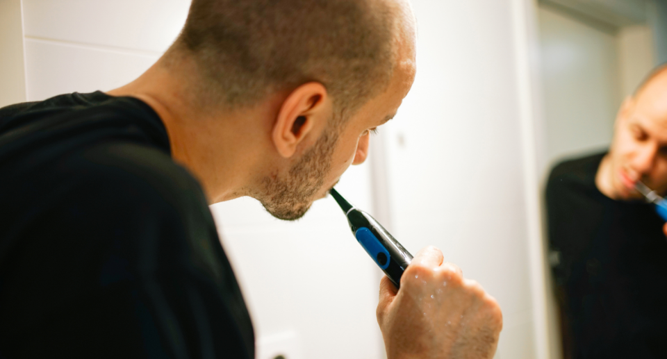 man brushing his teeth with an electric toothbrush in the mirror