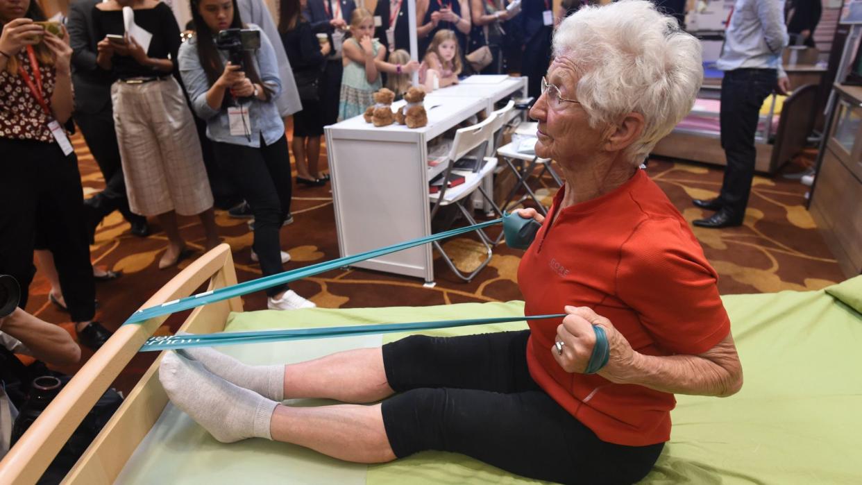  photo of older white woman with white hair and glasses using a resistance band as she warms up for gymnastics  