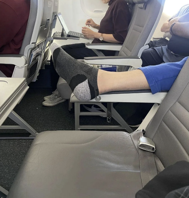 A person sitting on an airplane with their feet, in gray socks, resting on an armrest in front of them. Another passenger works on a laptop