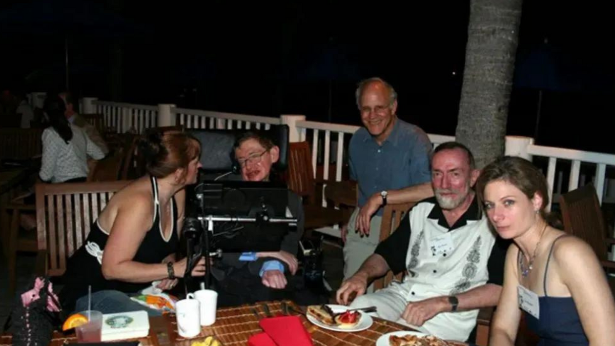 Five white people sit around a table. One person is in a wheelchair, which the person on the very left is looking at. The other three on the other side of the photo are smiling at the camera. 
