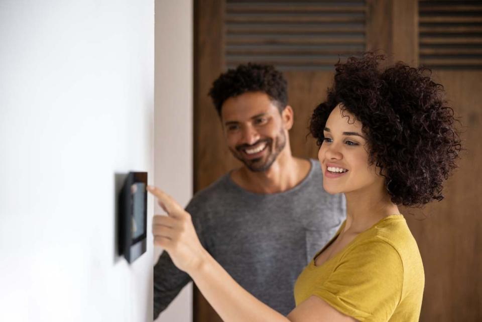 A man and a woman smile while she navigates the screen of a wall-mounted home security system.