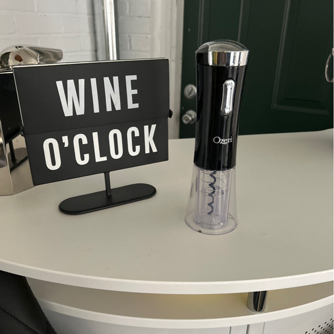 Electric wine opener next to a 'WINE O'CLOCK' sign on a table, suggesting a product for wine enthusiasts