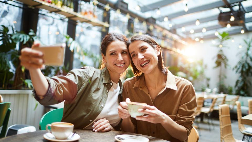 Happy attractive young women in casual shirts sitting at table in coffee shop and photographing together while drinking coffee.