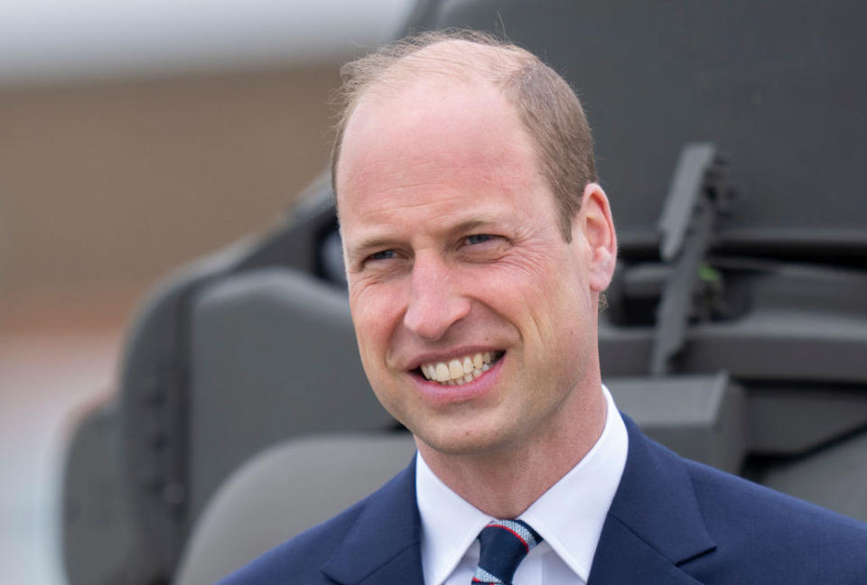 Prince William smiling, wearing a suit and tie, with a blurred background of what appears to be a military aircraft