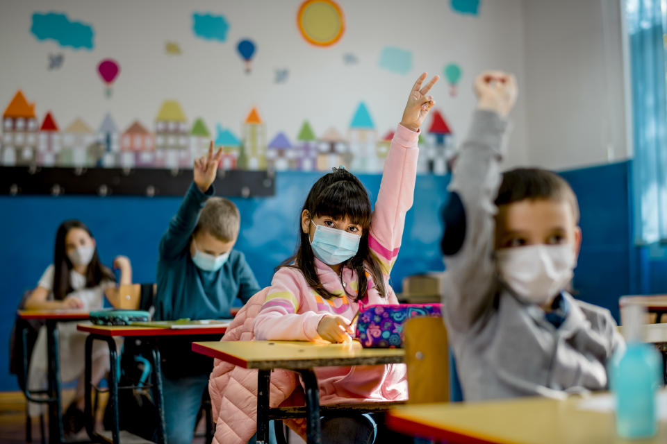 Elementary schoolchildren raise their hands while sitting at their desks in the classroom.