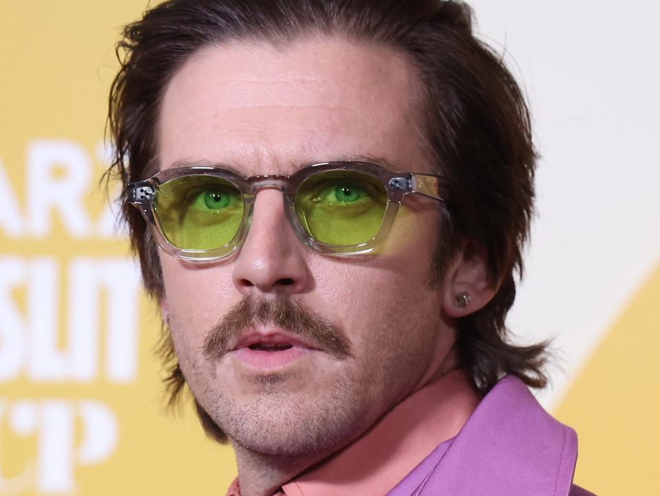 Dan Stevens poses for photos on the red carpet in a purple suit and green sunglasses.