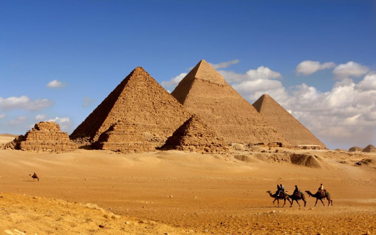 Three large pyramids are seen, with a number of smaller stepped pyramids around them and some camels with riders in the foreground