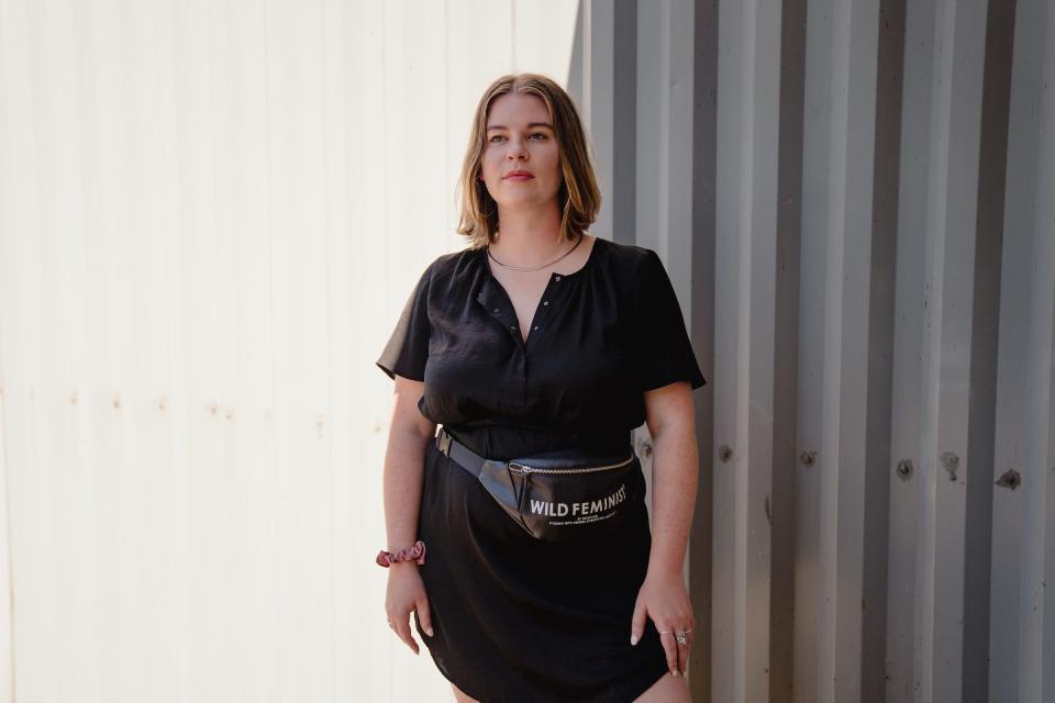 Tori Dunlap wearing a black short-sleeve top with hoop earrings, a necklace, and a fanny pack that says "Wild Feminist."