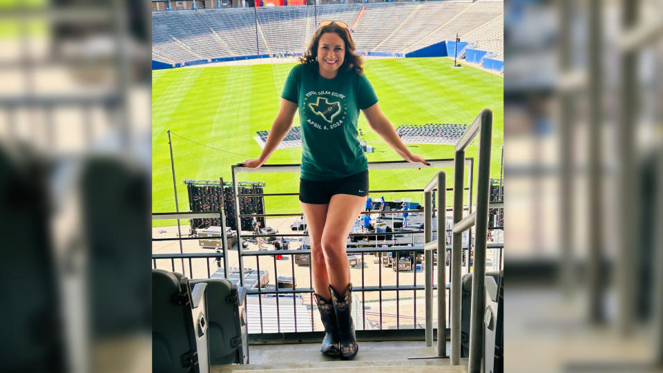 a woman with shoulder length brown hair stands on the steps of the stadium in the seating area and smiles at the camera.