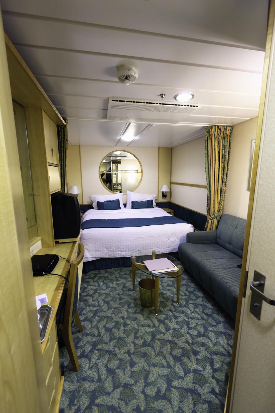 Interior view of a cozy, neatly arranged, nautical-themed bedroom with a double bed, a round mirror, a green sofa, a work desk, and an undressed window