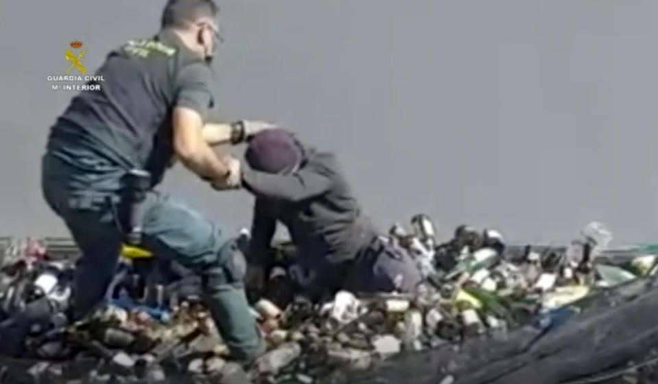 In this image taken from video made available by the Guardia Civil, an officer of the Guardia Civil helps a man out from under glass bottles in a container in Melilla, Spain, Friday Feb. 19, 2021. Spanish authorities say they have found and rescued 41 migrants who tried to reach continental Europe from North Africa in the past four days, some of them hiding inside a container of discarded glass bottles and a bag of toxic ash. (Guardia Civil via AP)