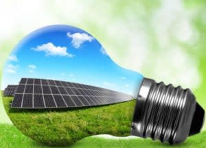 A lightbulb reflecting an image of solar panels in a grassy field under a blue, cloud-dotted sky; solar stocks