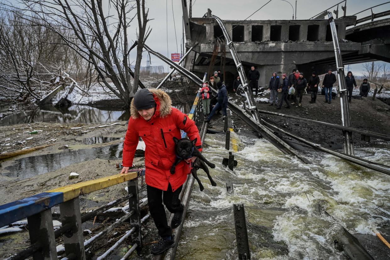 A man and his dog cross a river on a blown-up bridge.