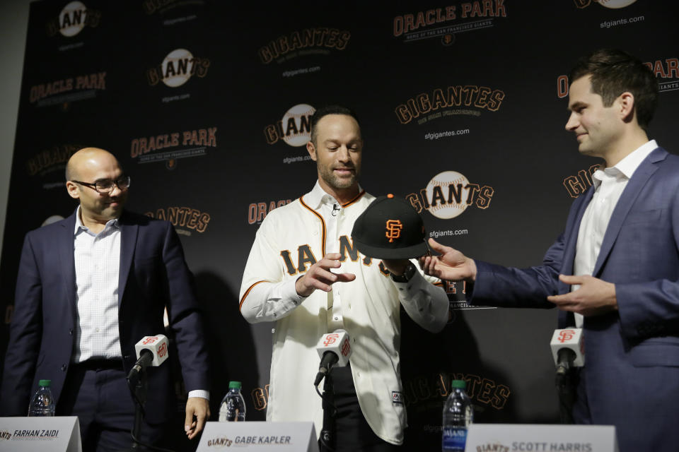 San Francisco Giants manager Gabe Kapler is given a new cap from general manager Scott Harris, right, as president of baseball operations Farhan Zaidi, left, looks on during a news conference at Oracle Park Wednesday, Nov. 13, 2019, in San Francisco. Kapler has been hired as manager of the San Francisco Giants, a month after being fired from the same job by the Philadelphia Phillies. Kapler replaces Bruce Bochy, who retired at the end of the season following 13 years and three championships with San Francisco. (AP Photo/Eric Risberg)