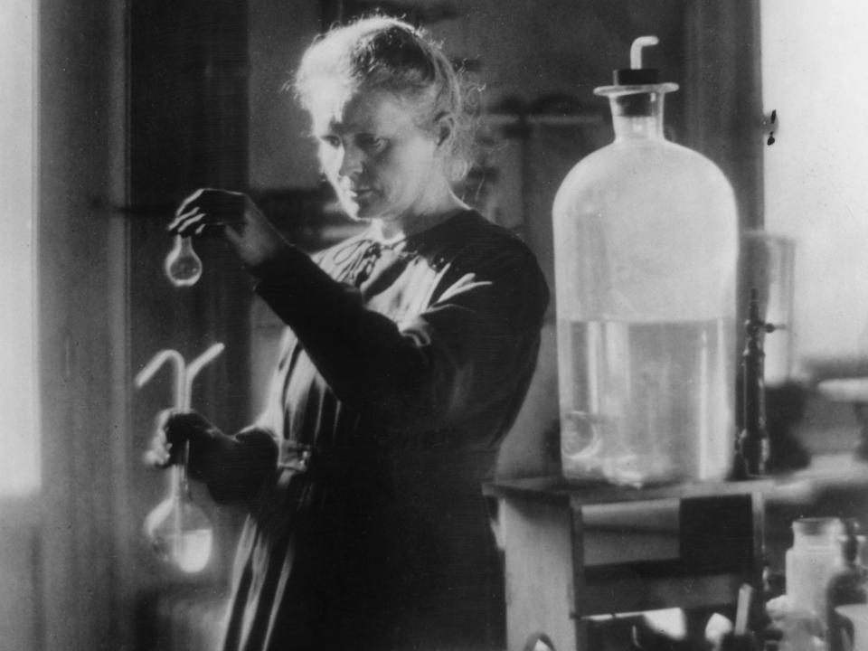 A black and white photo of Marie Curie holding up a glass jar for inspection while standing in a laboratory.