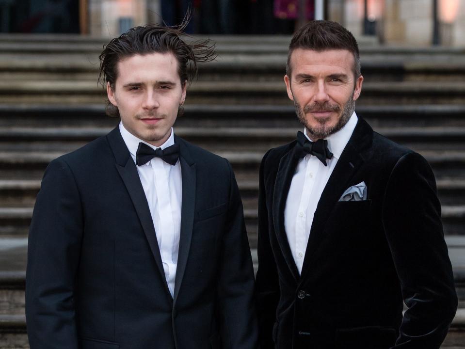 Brooklyn Beckham and David Beckham wear black suits as they're photographed on steps.