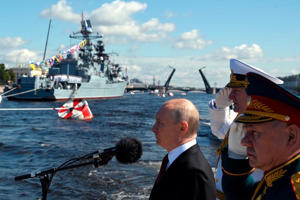Russian President Vladimir Putin stands in front of a microphone with a warship in the backrground