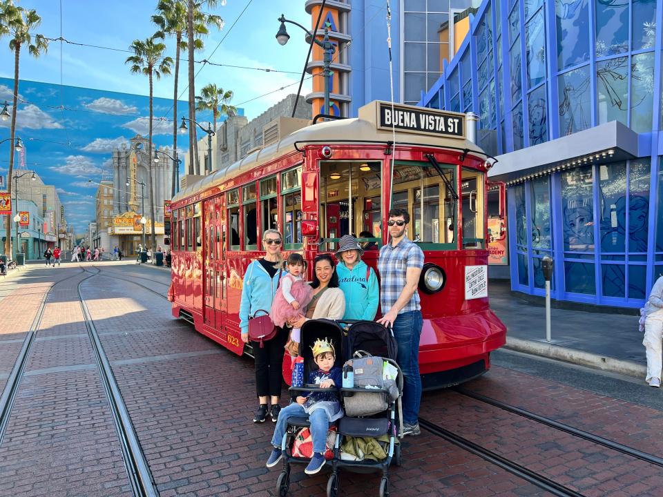 Three adults and two children in front of a red trolley with a sign that says "Buena Vista St" in California Adventure. One woman is holding a little girl, while a little boy sits in one side of a two-child stroller.