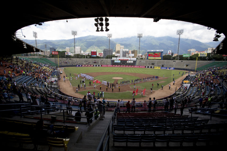 In this Oct. 12, 2018 photo, baseball players and fans stand for the national anthem during the opening season baseball game between Leones de Caracas and Tiburones de la Guaira in Caracas, Venezuela. For the second straight year, state-run oil company PDVSA had to step in with a $12 million lifeline to pay for everything from imported baseballs to the salaries of seven foreign-born players _ most of them minor league prospects from the U.S. _ on each team's roster. (AP Photo/Fernando Llano)