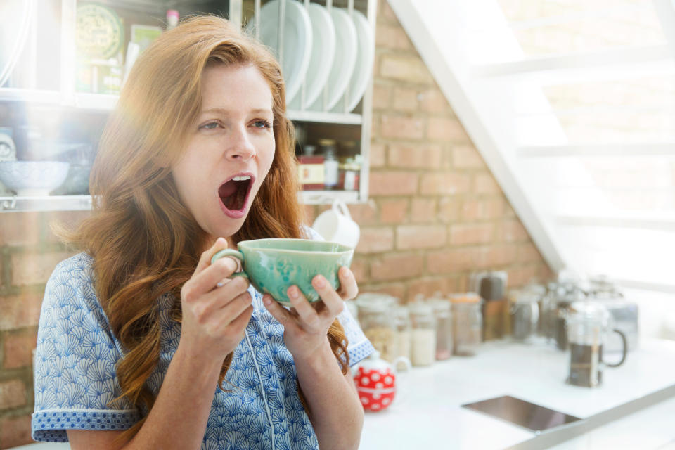 A woman with red hair yawns while holding a mug with both hands