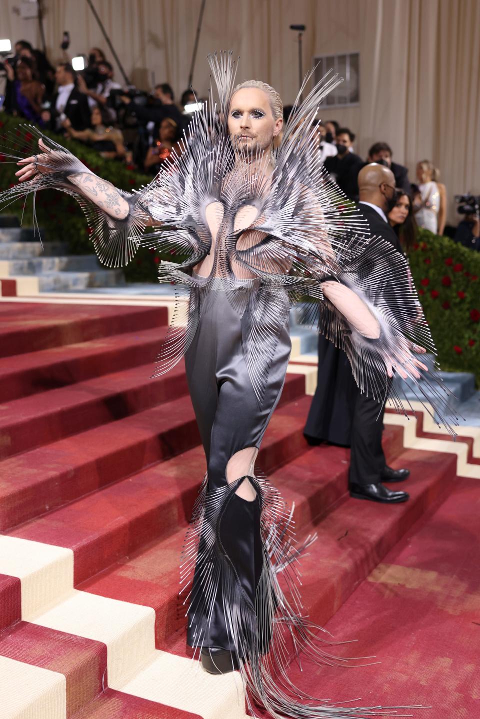 Fredrik in an ombre metallic sculptural piece with sharp thin pieces of material coming out around his shoulders, arms, and legs. He's wearing metallic ombre pants but his torso and arms are expose besides the spiky pieces attached to a sheer underlay fabric.